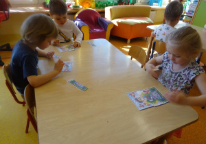 Troje dzieci siedzi przy stole i składa z części obrazek łąki.